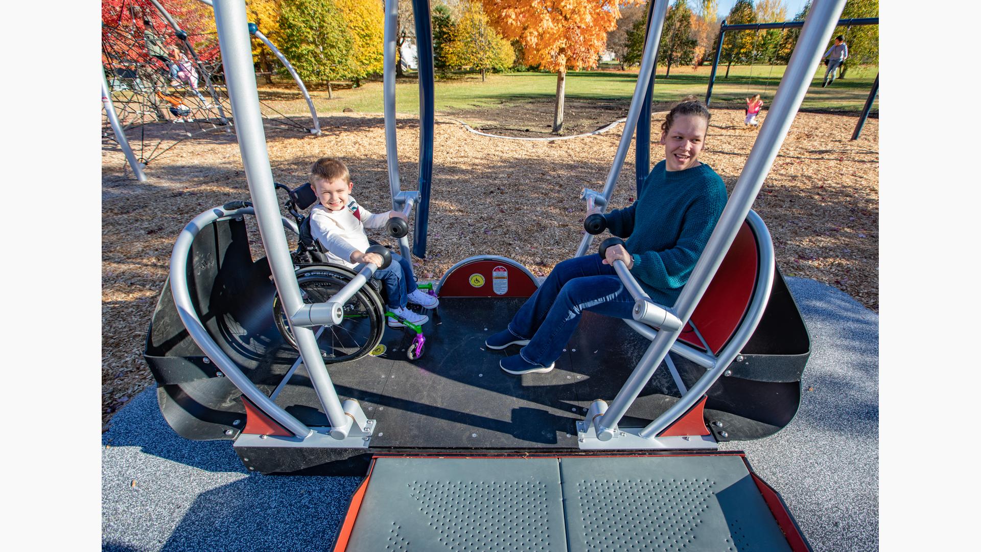 A mom and her son in his wheelchair are happy to swing together on the accessible swing.