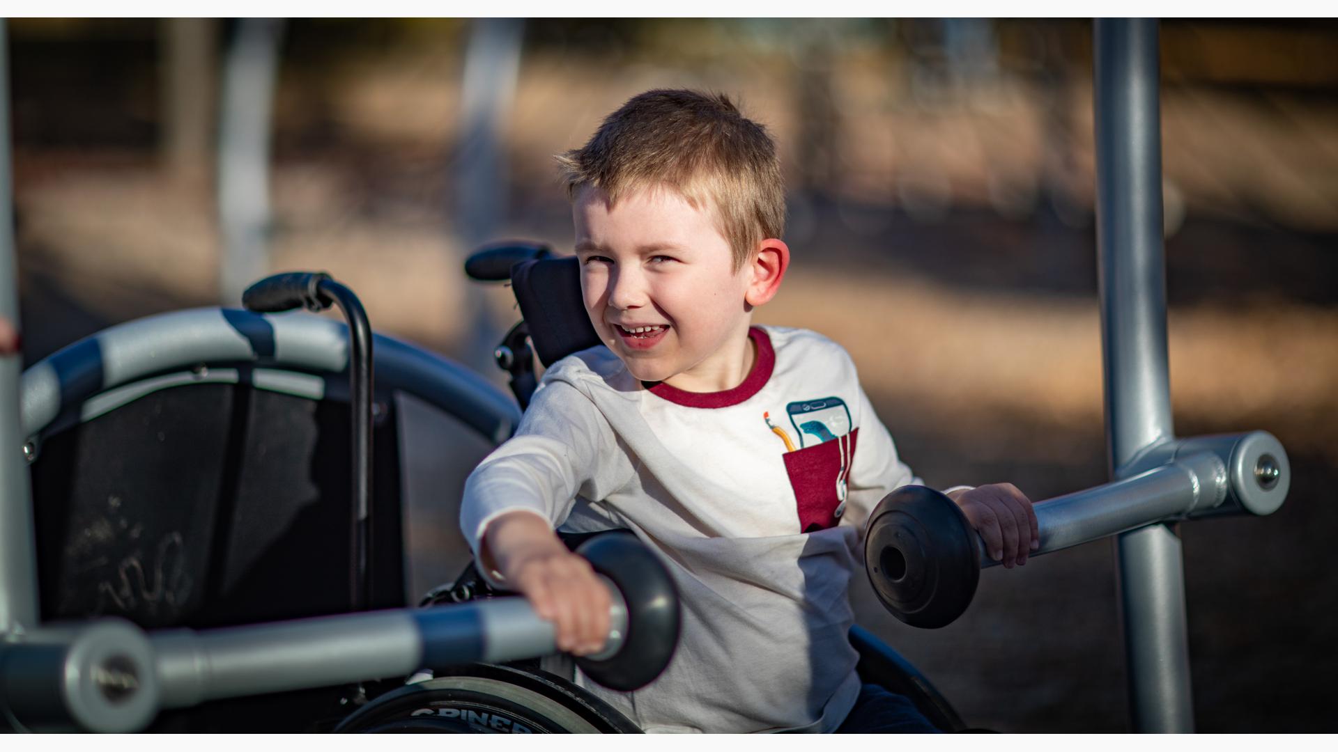 An excited boy in his wheelchair is backed into the accessible swing bay and holds onto the handlebars.